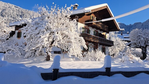 Eine Holzhütte mit Balkon, umgeben von schneebedeckten Bäumen und einem Bergpanorama.