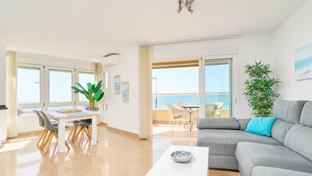 Ein modernes Wohnzimmer mit grauem Sofa, einem Esstisch mit Stühlen, einem großen Fenster und einem Balkon mit Meerblick.