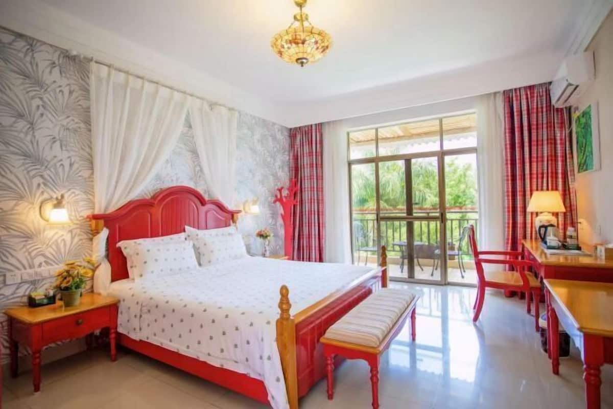 A hotel room with a bed, a desk, a chair, a bench, a nightstand, and a window with a view of greenery.