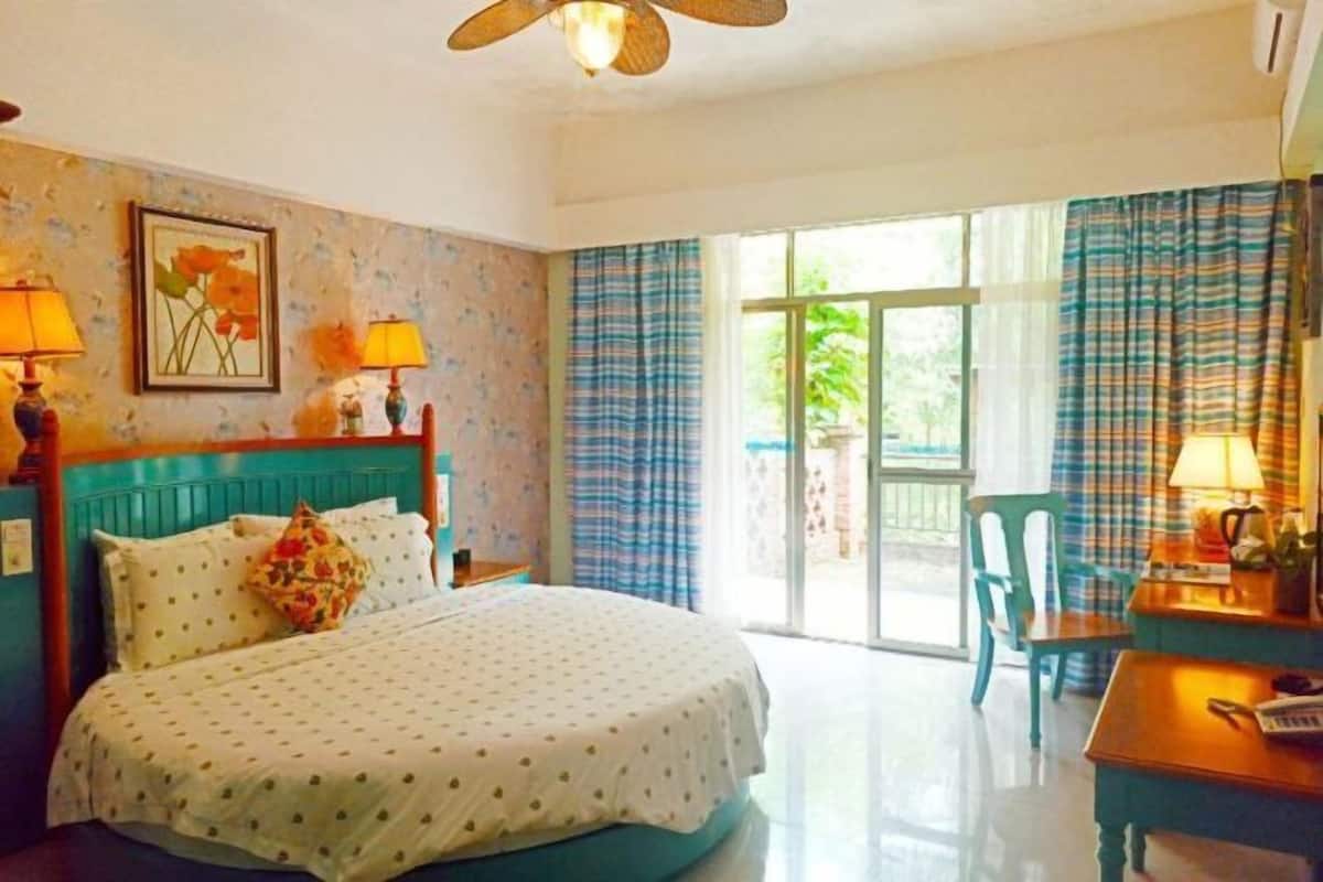 A hotel room with a bed, a desk, a chair, a window with curtains, and a ceiling fan.