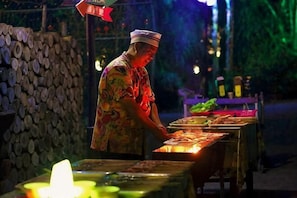 A person preparing food at an outdoor market stall with a table, a stack of wooden logs, and a night-time setting. - Forest Inn (Hainan University, Zhangzhou Botanical Garden Thermoscience Academy) (Danzhou)