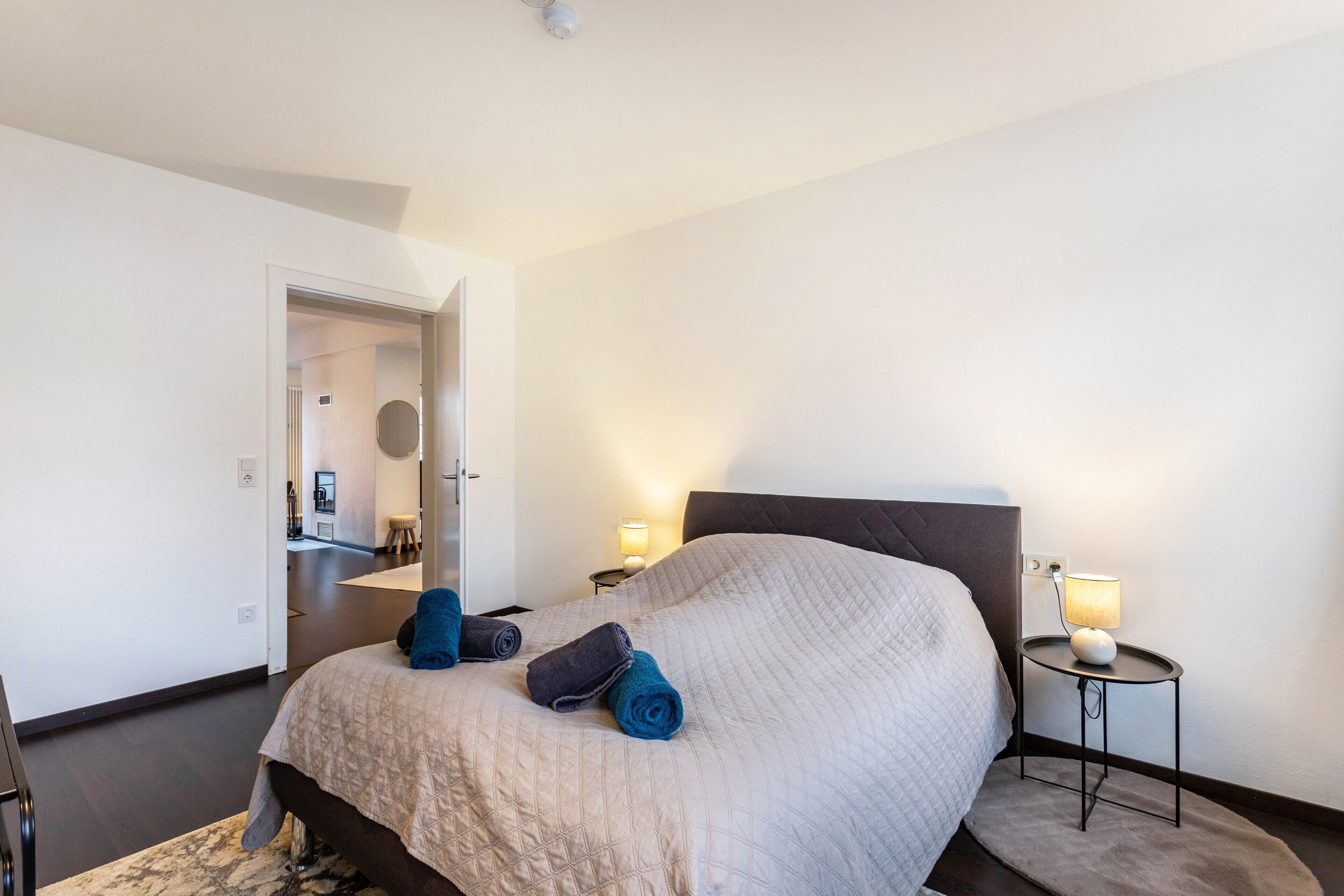 A hotel room with a bed, two bedside lamps, and a door leading to another room.