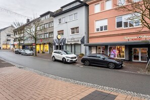 Une vue de rue avec des voitures garées, une horloge et des devantures de magasins, notamment une bijouterie et un restaurant.