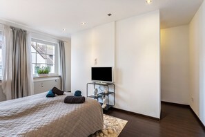 Une chambre à coucher avec un lit, un téléviseur posé sur un meuble, une fenêtre avec des rideaux et un radiateur.