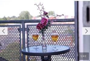 Et rundt bord med to glas med amberfarvet væske, en vase med lilla blomster og et metalhegn i baggrunden.