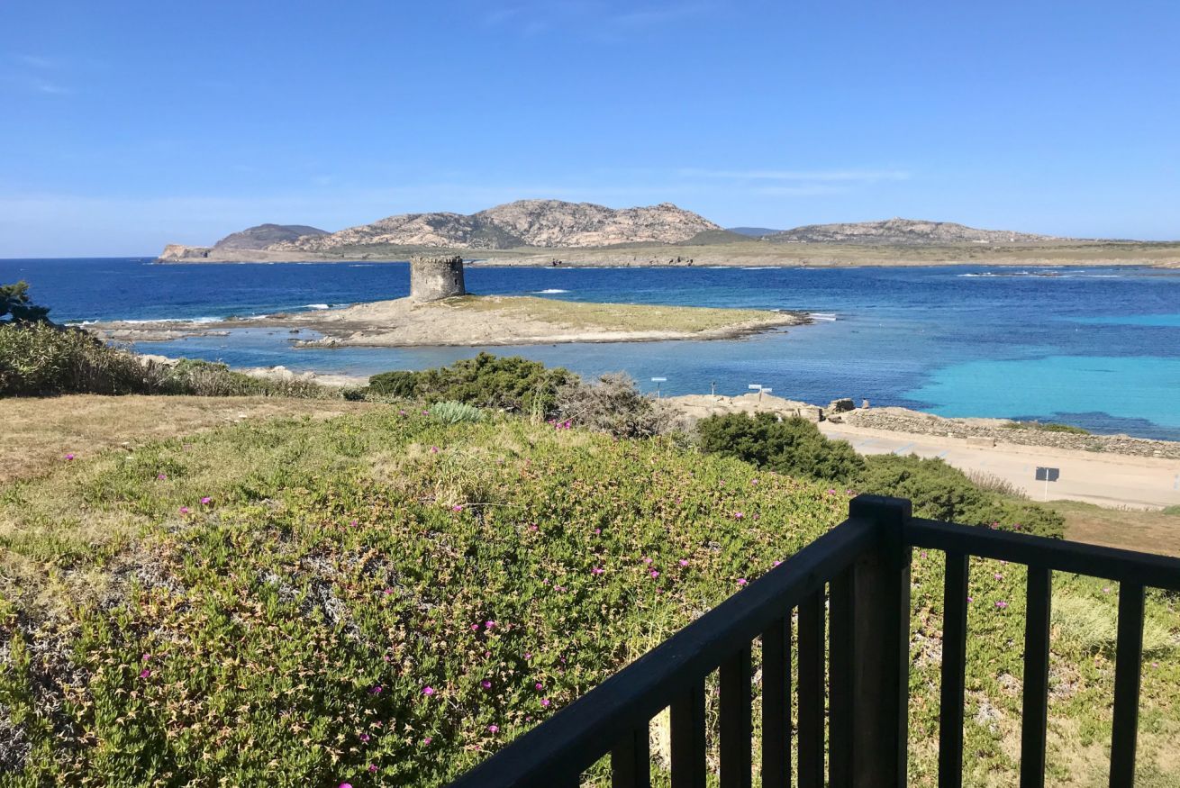 A coastal landscape with a stone structure, clear blue sea, and distant hills.