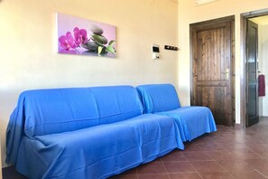 A room with a blue sofa, a wooden door, and a wall-mounted thermostat. - Charming 19 sq m studio apartment.It features a fully equipped kitchenette, a double bed or two single beds, and a private bathroom with shower, bidet, and hairdryer. The apartment also boasts an outdoor space with a sea view, a patio or terrace, and a t (Stintino)