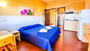 A bedroom with a bed, a small table, and a nightstand. There is a kitchen area with cabinets and a refrigerator. - Charming 19 sq m studio apartment.It features a fully equipped kitchenette, a double bed or two single beds, and a private bathroom with shower, bidet, and hairdryer. The apartment also boasts an outdoor space with a sea view, a patio or terrace, and a t (Stintino)