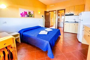 A bedroom with a bed, a small table, and a nightstand. There is a kitchen area with cabinets and a refrigerator. - Charming 19 sq m studio apartment.It features a fully equipped kitchenette, a double bed or two single beds, and a private bathroom with shower, bidet, and hairdryer. The apartment also boasts an outdoor space with a sea view, a patio or terrace, and a t (Stintino)