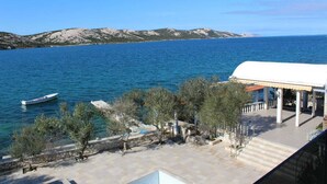 Un hotel a la costa amb terrassa que domina el mar, una embarcació amarrada a prop i un paisatge de muntanyes al fons.