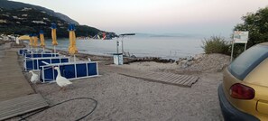 A beachside area with blue and yellow umbrellas, a wooden deck, and a yellow car parked nearby. - Aenaon Corfu Luxury Apartments 05 - Athena Apt (Corfu)
