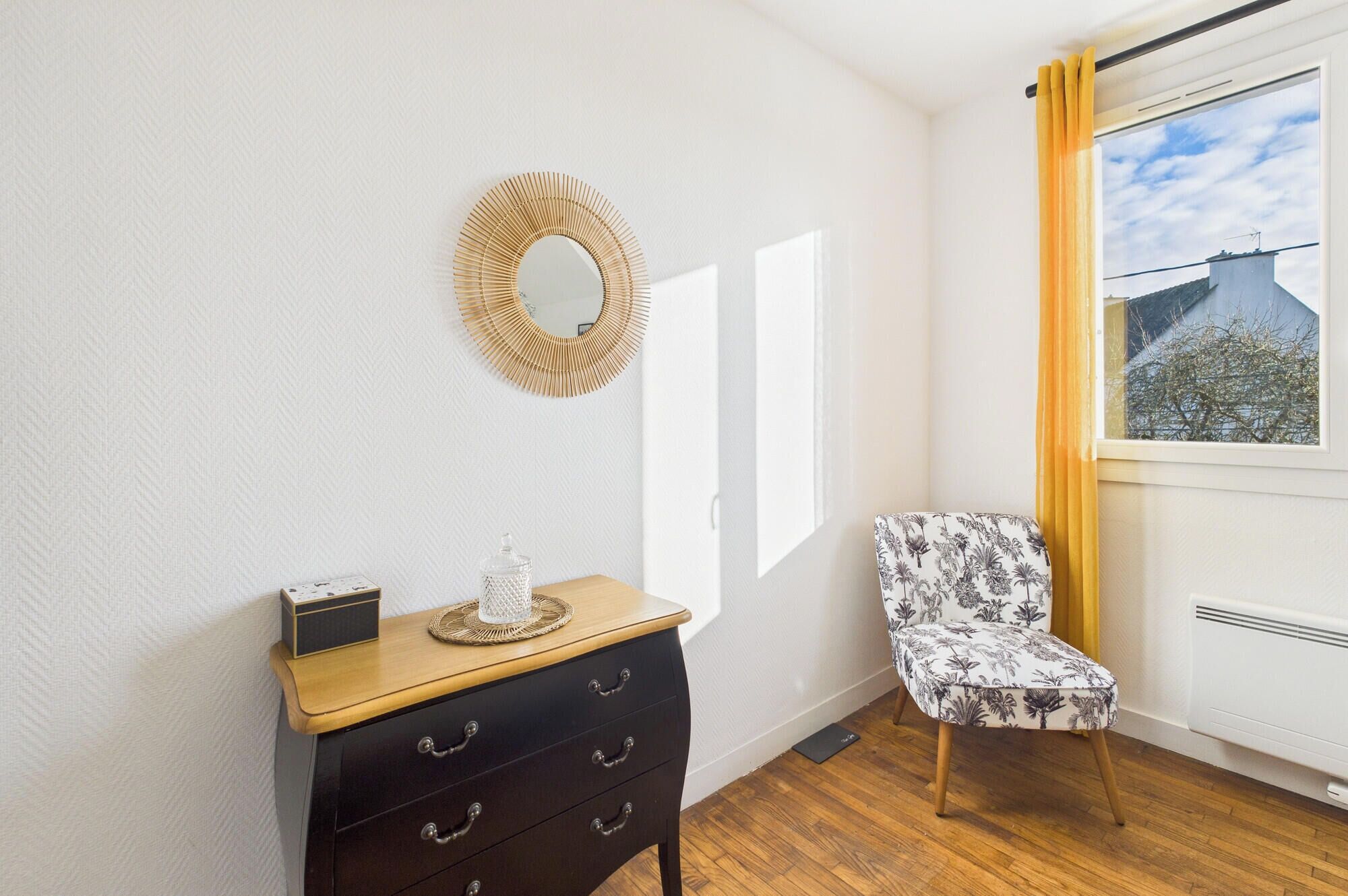 Een kamer met een houten dressoir, een gestreept fauteuil, een spiegel aan de muur en een raam met gordijnen.