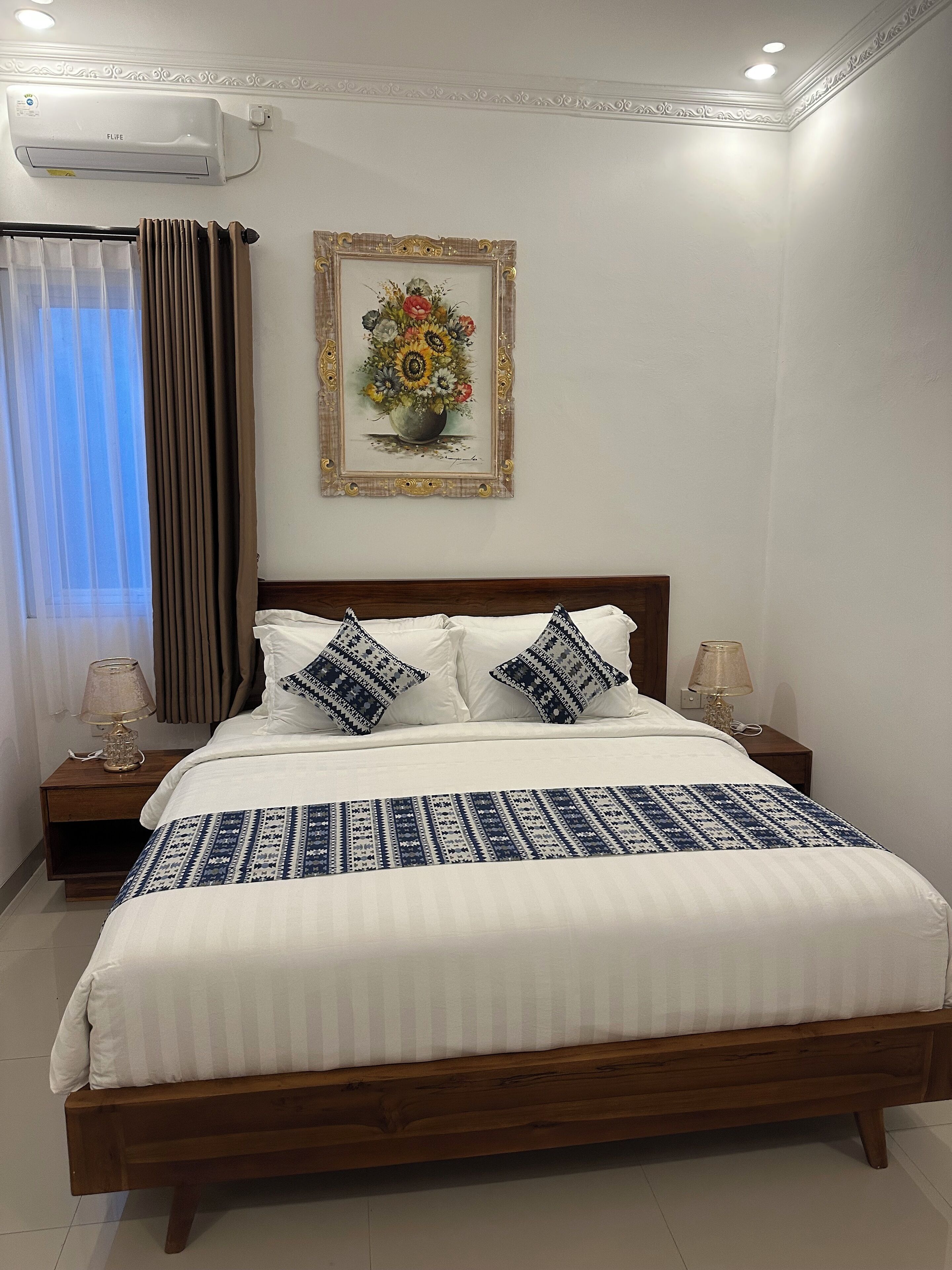 A neatly made bed with white bedding and patterned pillows, a wooden headboard, and a framed floral artwork above it.