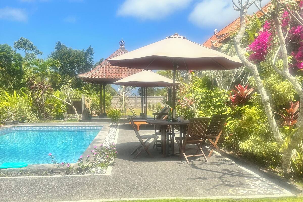 A swimming pool with a tiled edge, surrounded by lush greenery and a gazebo with a tiled roof.