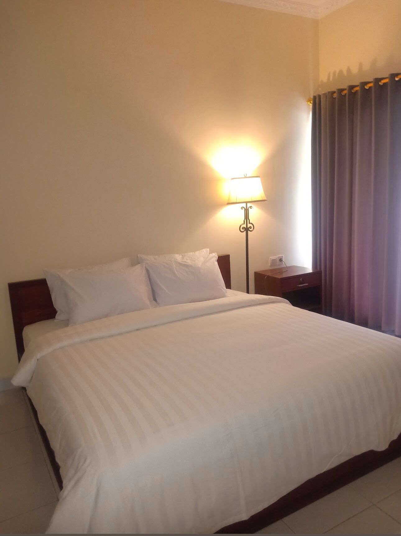 A neatly made bed with white linens, a bedside table with a lamp, and a window with curtains.