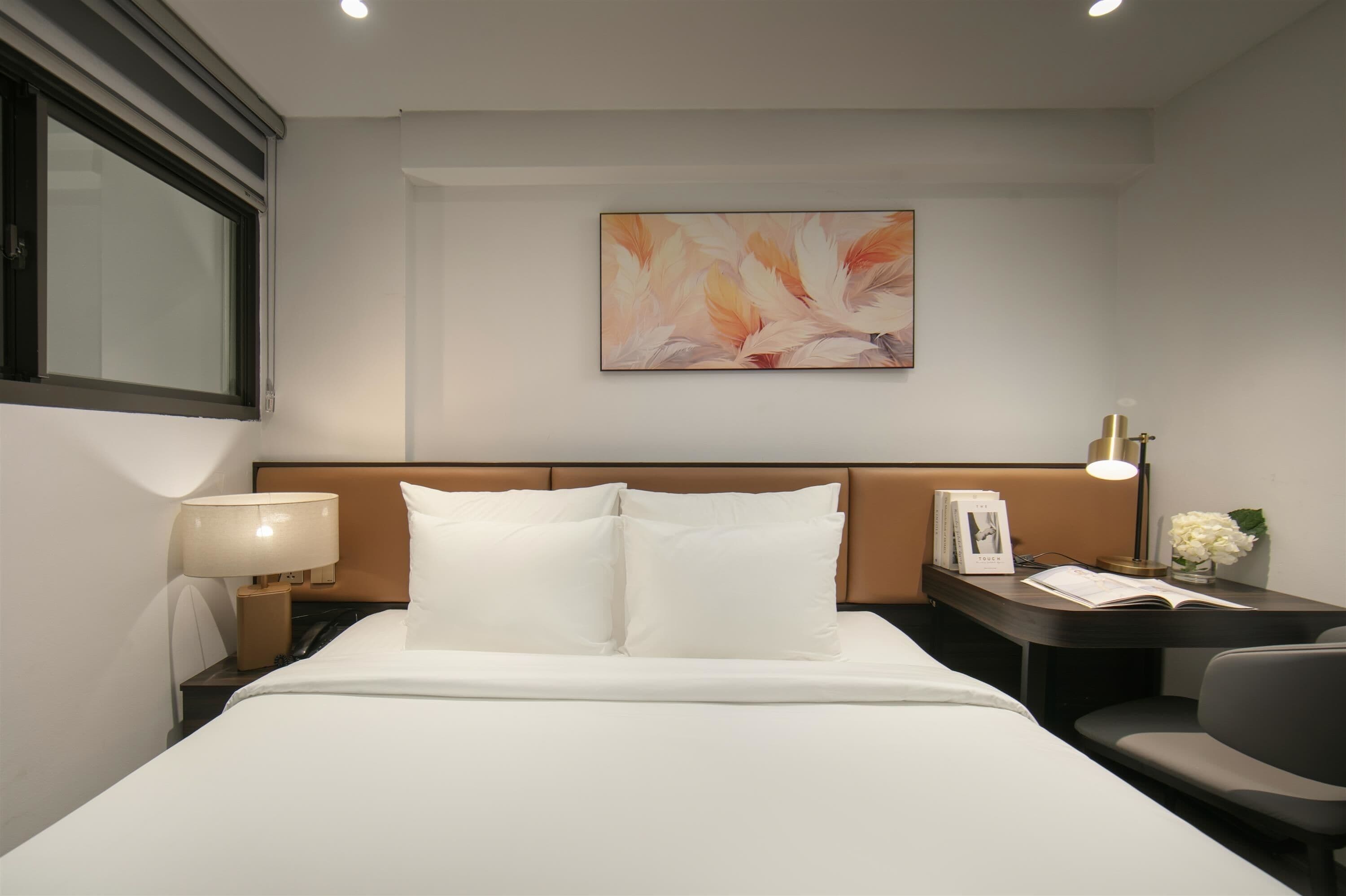 A hotel room with a bed, a nightstand with a lamp, a bedside table with a book and a vase of flowers, and a wall-mounted painting.