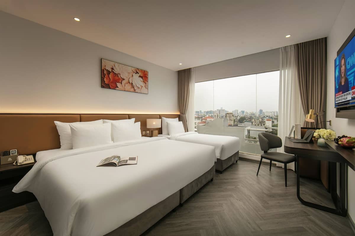 A hotel room with two beds, a desk, a chair, a TV, and a large window with a city view.