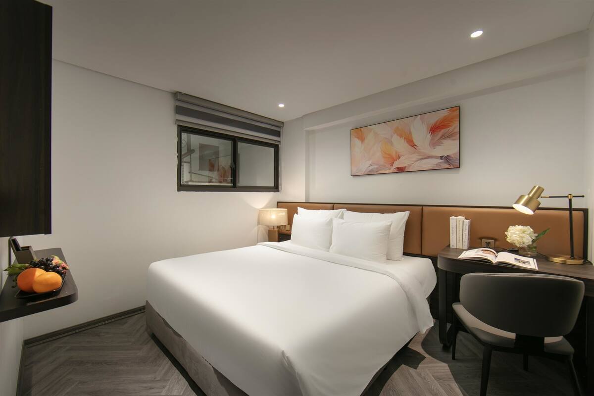 A hotel room with a large bed, a desk with a chair, a nightstand with a lamp, and a wall-mounted artwork.
