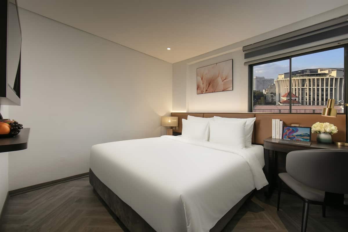 A hotel room with a large bed, a desk with a chair, a window with a city view, and a wall-mounted artwork.