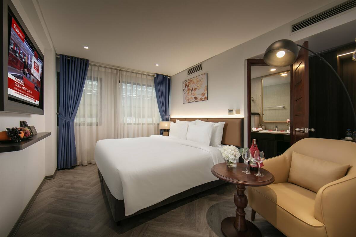 A hotel room with a large bed, a sofa, a small table with glasses, and a TV on the wall.