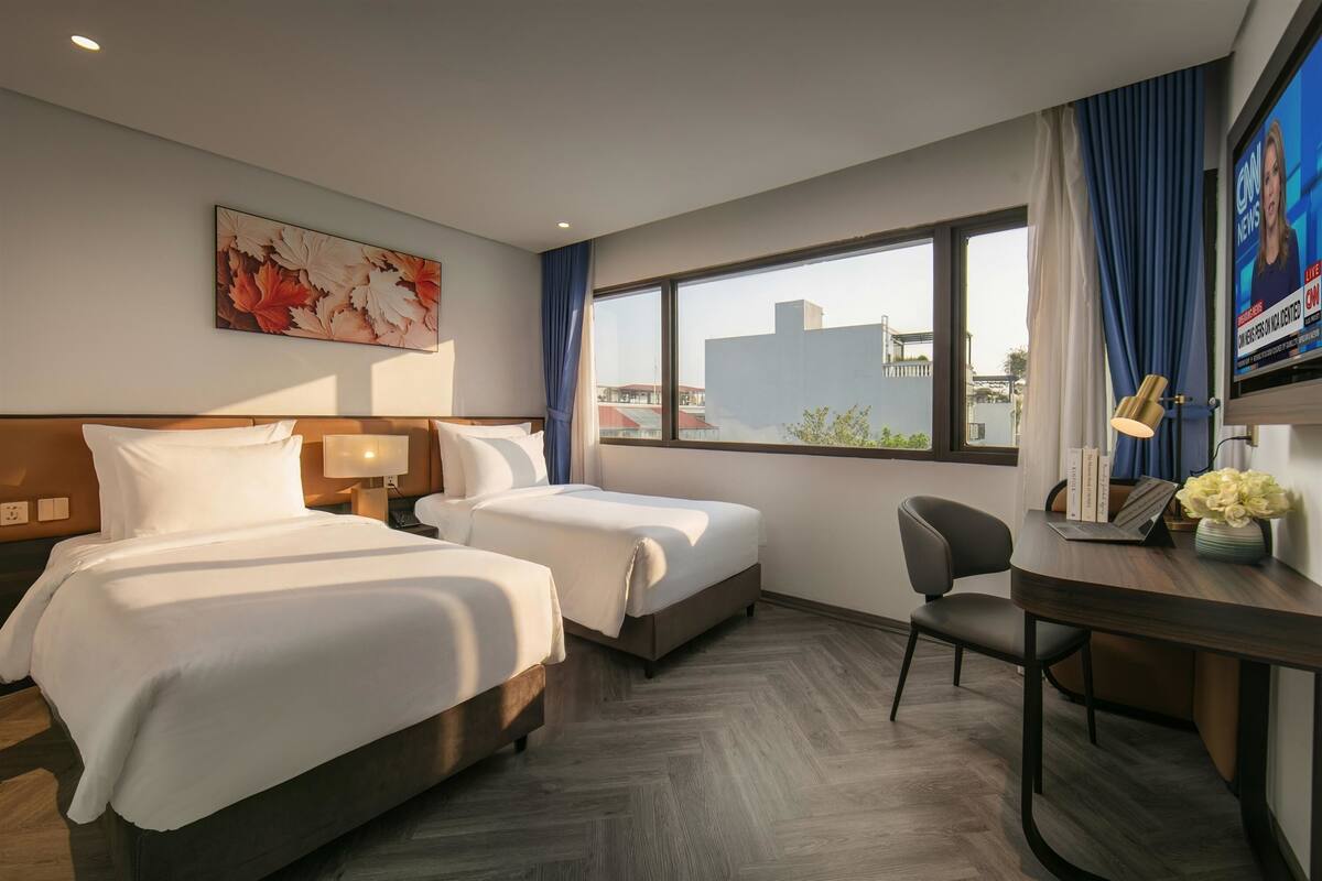 A hotel room with two beds, a desk with a chair, a TV, and a window with a view of buildings.