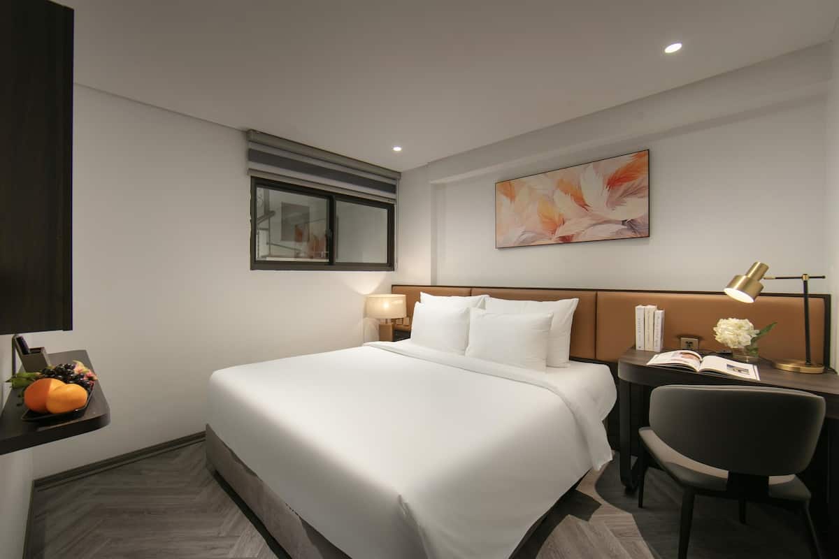 A hotel room with a large bed, a desk with a chair, a nightstand with a lamp, and a wall-mounted artwork.