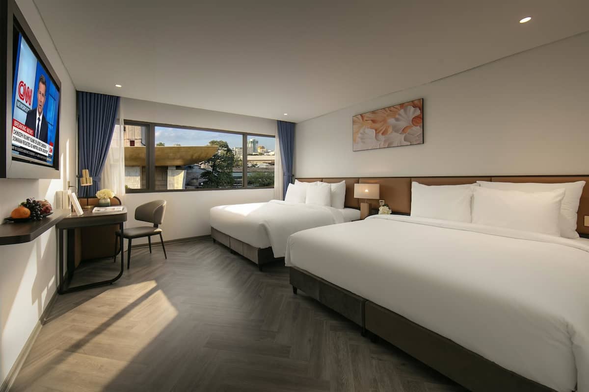 A hotel room with two beds, a desk with a chair, a TV mounted on the wall, and a window with a view of the city.