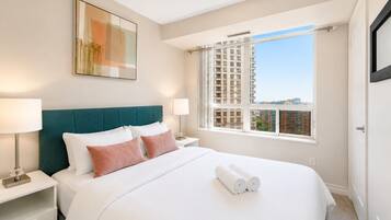 A hotel room with a bed, two bedside tables, a window with a city view, and a painting on the wall.
