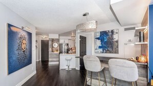 A modern kitchen with white cabinets, a stainless steel refrigerator, and a bar area with two white bar stools. - Luxury 2BR+Den | Steps from SQ1 | Panoramic Views! (Mississauga)