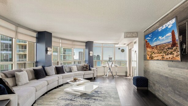 A modern living room with a large sectional sofa, a coffee table, and a large window offering a city view. - Luxury 2BR+Den | Steps from SQ1 | Panoramic Views! (Mississauga)