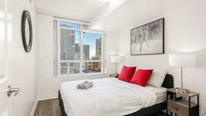 Et moderne soveværelse med et stort vindue, en seng med hvidt sengetøj og røde puder, et natbord med en lampe og udsigt over bybilledet.