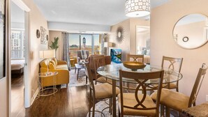 Una sala de estar moderna con una mesa de comedor de vidrio, sofá amarillo y vistas a un paisaje urbano a través de amplias ventanas.