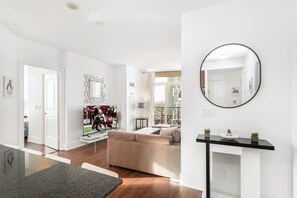 A modern living room with a flat-screen TV, a beige sofa, a dining area with a glass table, and a large window offering a city view.
