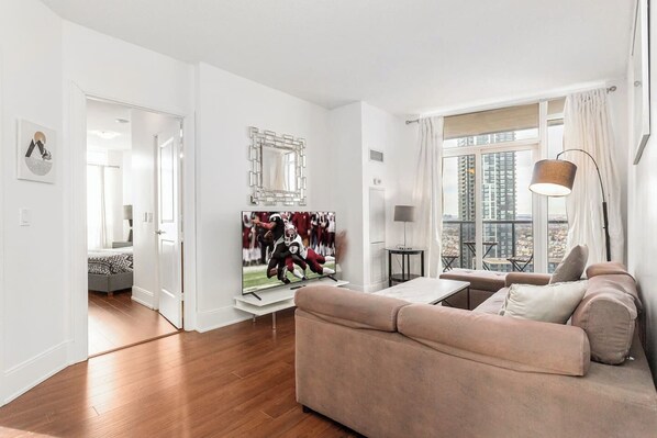 A modern living room with a large flat-screen TV, a beige sofa, a small coffee table, and a view of a cityscape through large windows.