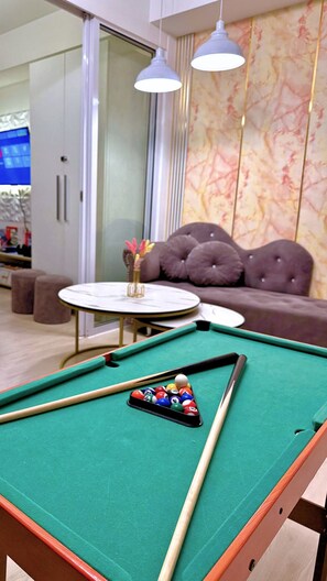 A pool table with billiard balls and cues, a sofa, and a small table with a vase of flowers in a room with a marble wall and pendant lights.