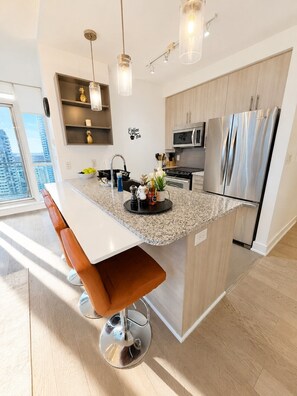 A modern kitchen with a central island, stainless steel appliances, and a view of the city. - Penthouse 2BR+Den | Skyline Views • SQ1 • Parking (Mississauga)