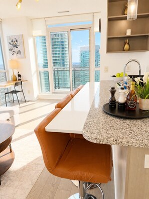 A modern kitchen with a granite countertop, a bar stool, a dining table, a window with a city view, and a shelf with kitchen items. - Penthouse 2BR+Den | Skyline Views • SQ1 • Parking (Mississauga)