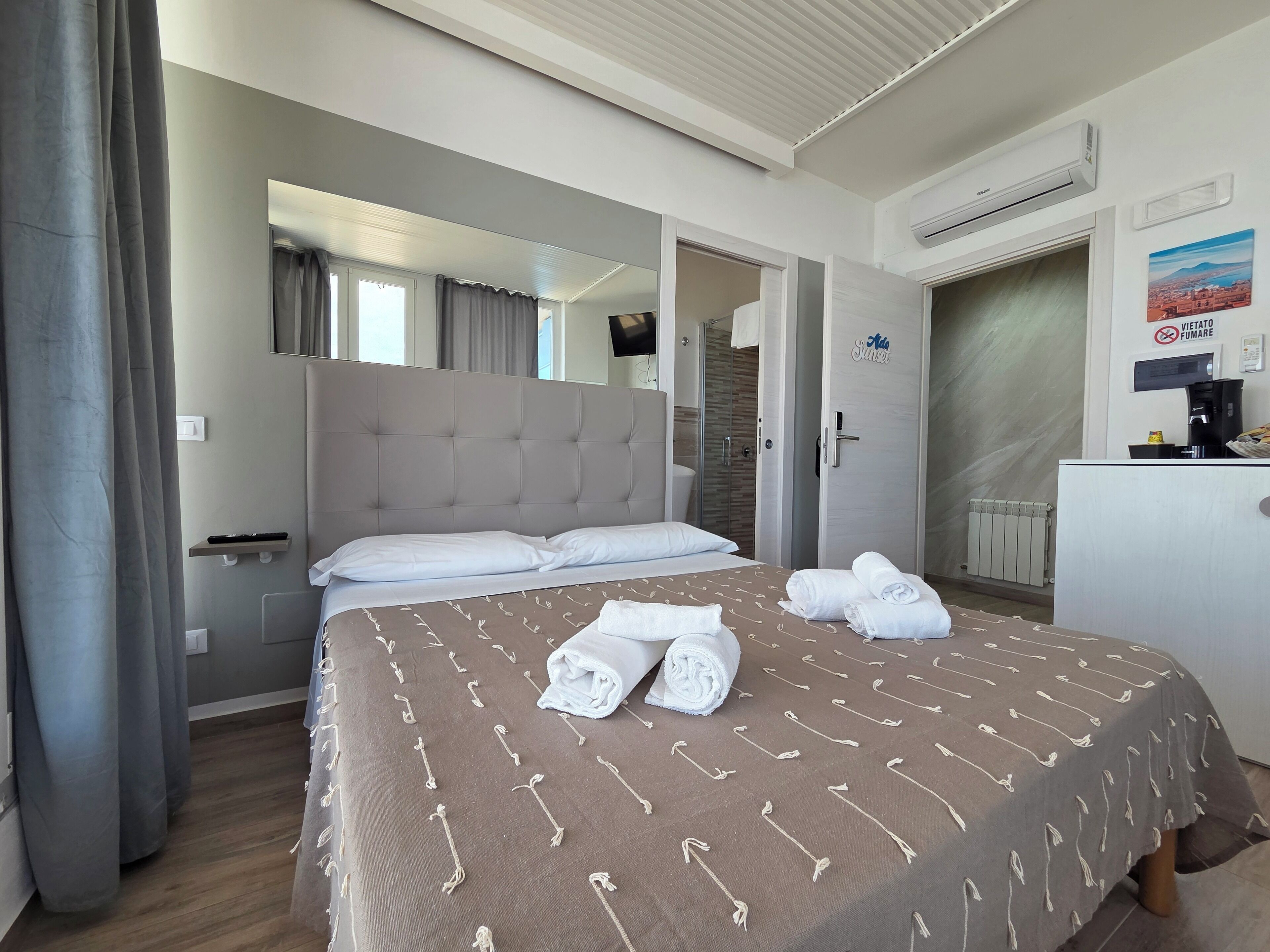 A hotel room with a large bed, a mirror, a wardrobe, a bathroom, and a desk with a coffee machine.