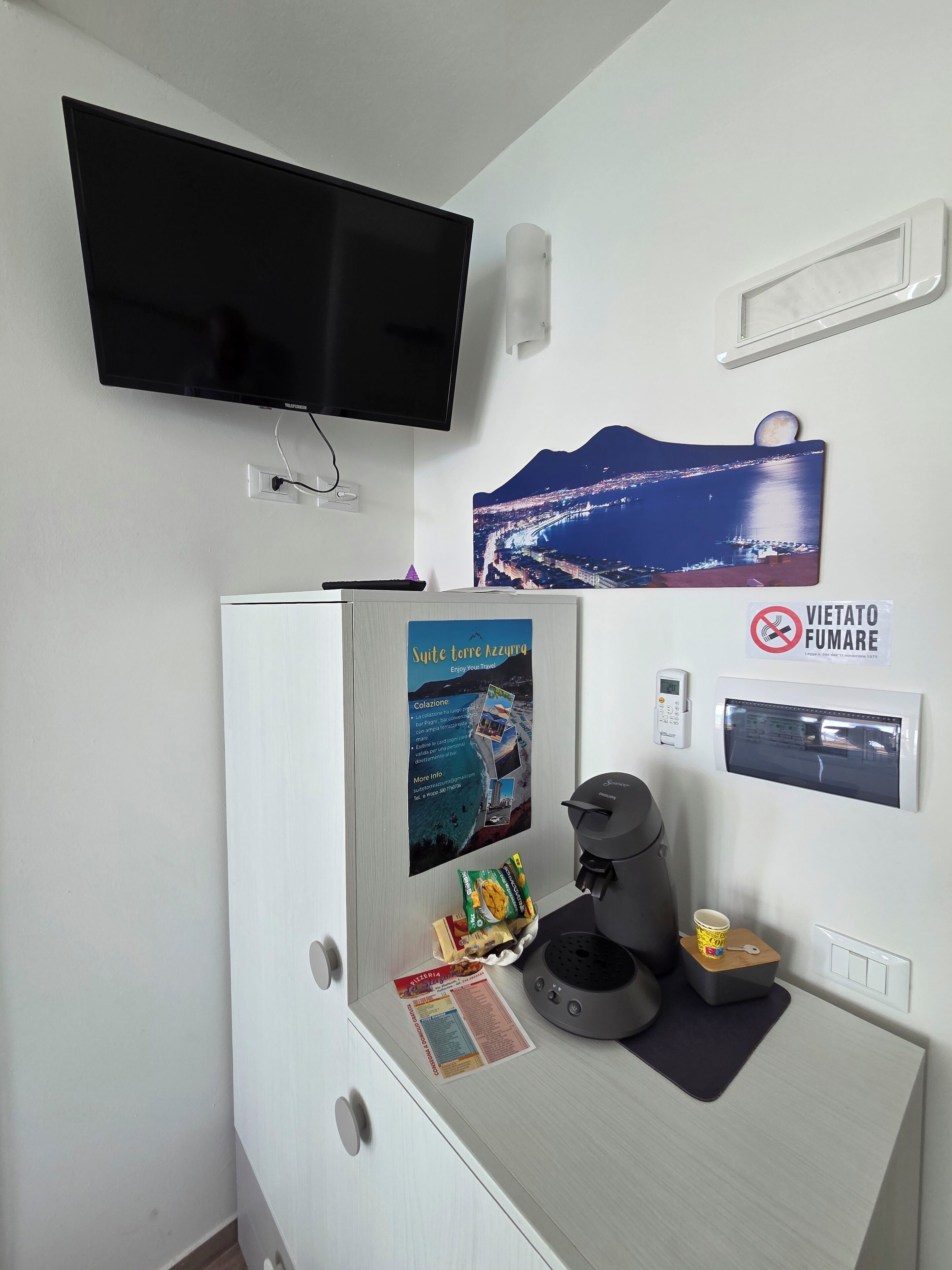 Une chambre d’hôtel équipée d’un téléviseur à écran plat, d’une affiche représentant une ville côtière, d’un panneau « interdiction de fumer », d’une machine à café et d’une petite table avec une tasse et un paquet de collations.