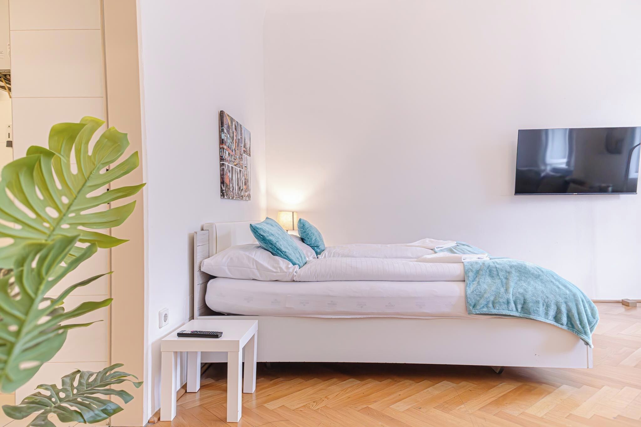 Ein modernes Schlafzimmer mit einem Bett, einem kleinen Nachttisch, einem an der Wand montierten Fernseher und einer großen Pflanze.