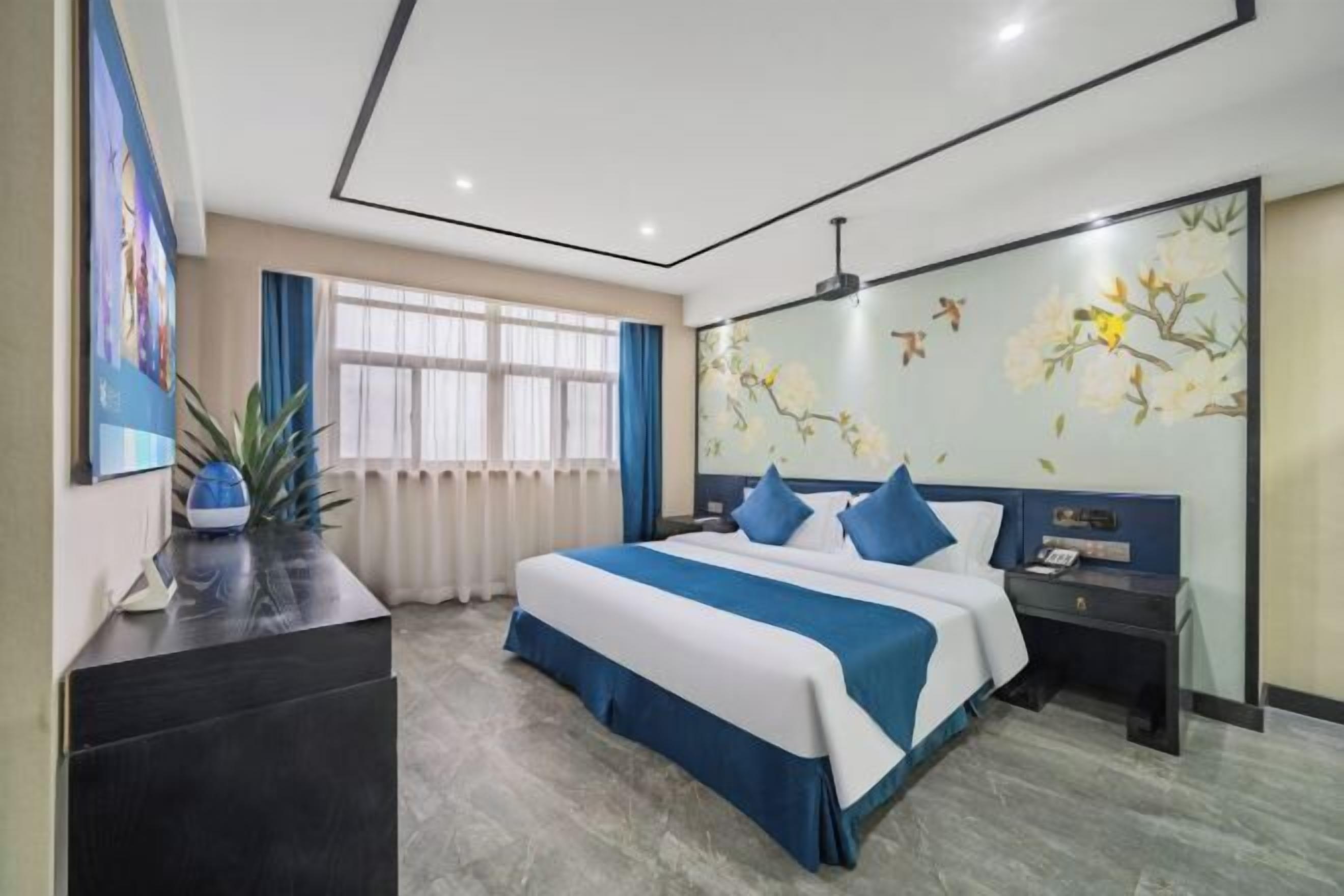 A hotel room with a large bed, a bedside table, a nightstand, a wall-mounted TV, and a window with curtains.