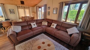 Una sala de estar moderna con un sofá seccional de cuero marrón, una mesa de centro redonda de madera con detalles naranja y ventanas amplias que ofrecen vistas a la vegetación exterior.