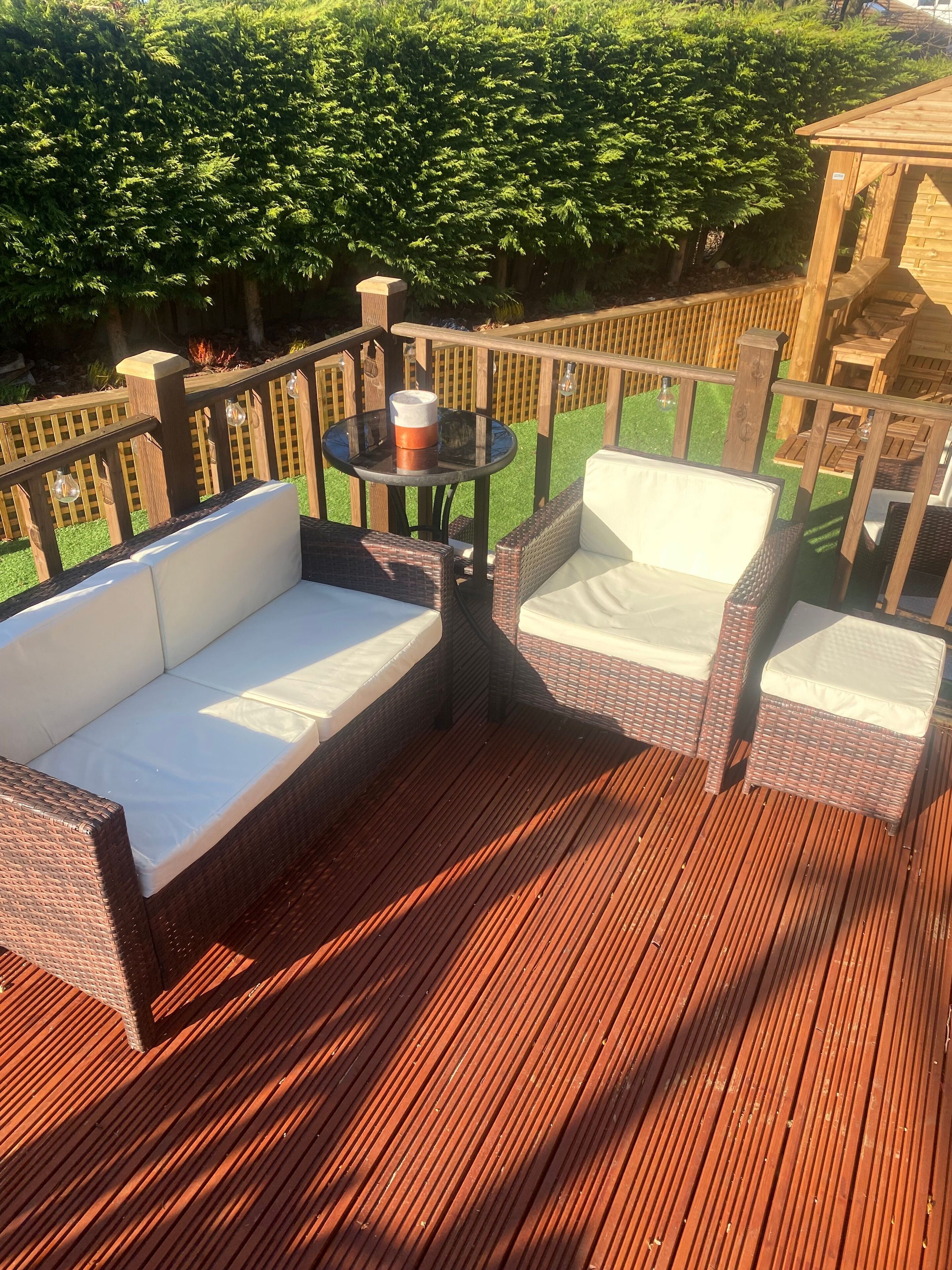 Une terrasse en bois, agrémentée de meubles en osier, notamment d’un canapé et de fauteuils, ainsi qu’une petite table ronde sur laquelle se trouve une tasse.