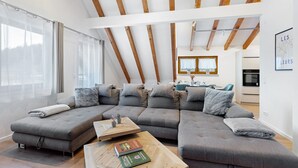 Ein modernes Wohnzimmer mit einem Ecksofa, Holzbalken und einem Essbereich im Hintergrund.