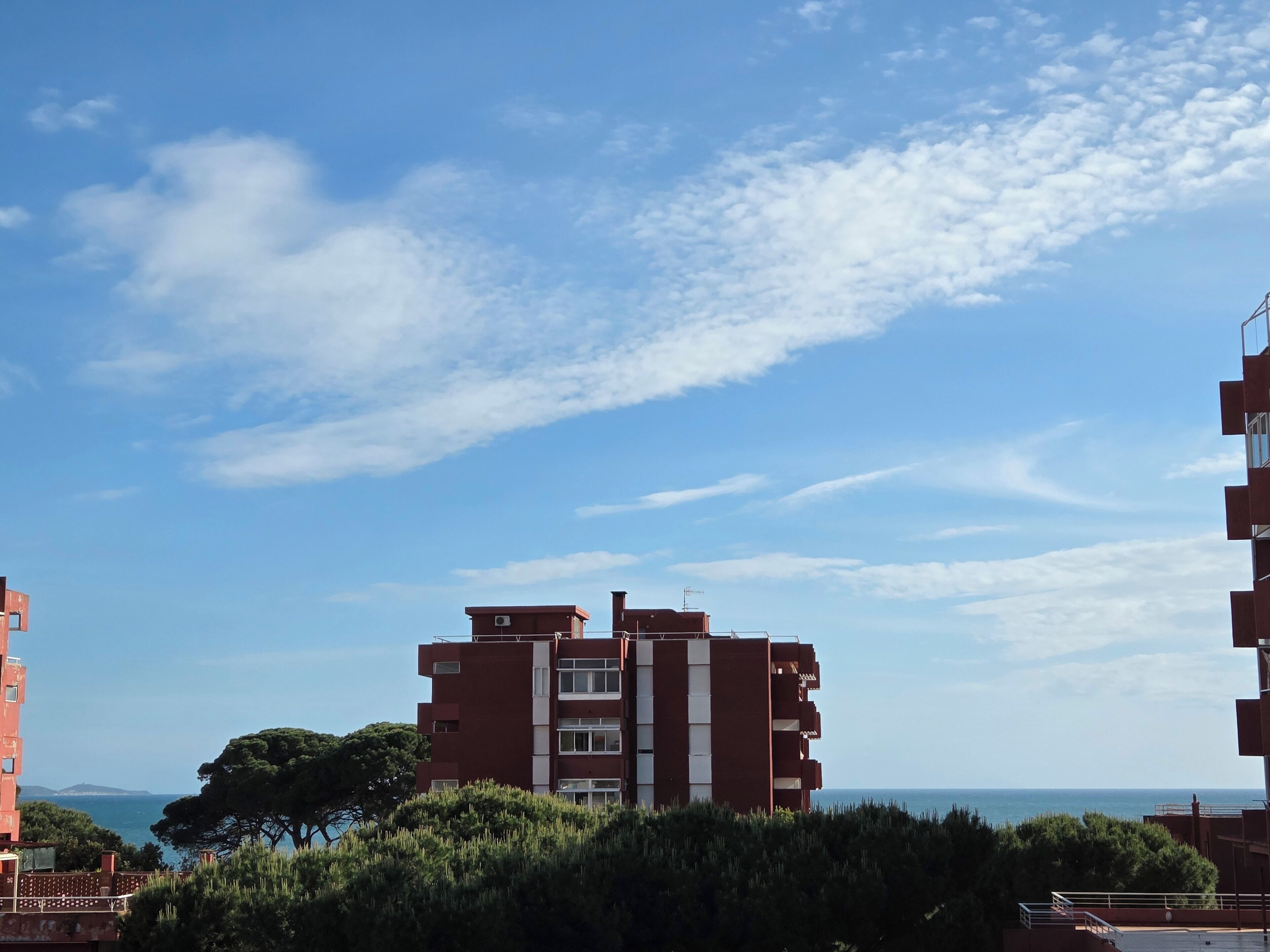 Un edificio de varios pisos con fachada roja y detalles blancos, rodeado de vegetación y con vista a un cuerpo de agua.