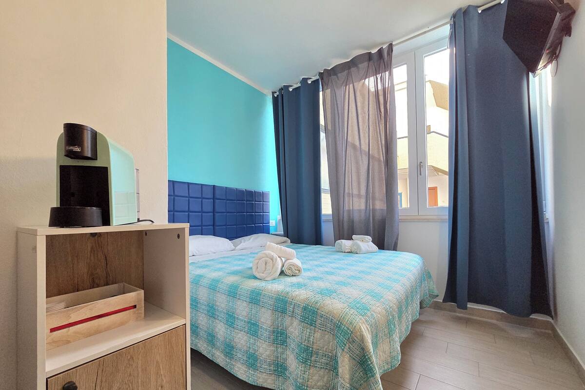 Una habitación de hotel con pared azul, cama con colcha a cuadros, mesita de noche con cafetera y ventana con cortinas.