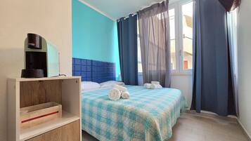 A hotel room with a blue wall, a bed with a checkered bedspread, a bedside table with a coffee maker, and a window with curtains.