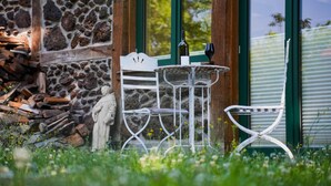 Un edificio de piedra con puerta de cristal, una mesa de metal blanco con sillas, y una estatua al frente.