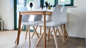 Un comedor moderno con una mesa de madera y sillas blancas, ventanas amplias con vista a follaje, y un jarrón con flores sobre la mesa.