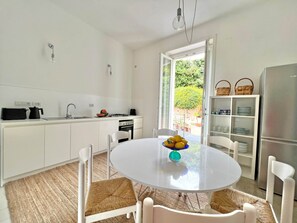 A modern kitchen with white cabinetry, a dining table with chairs, a refrigerator, and a view of greenery outside. - Villa Ipomea - Cielo & Terra (Salerno)
